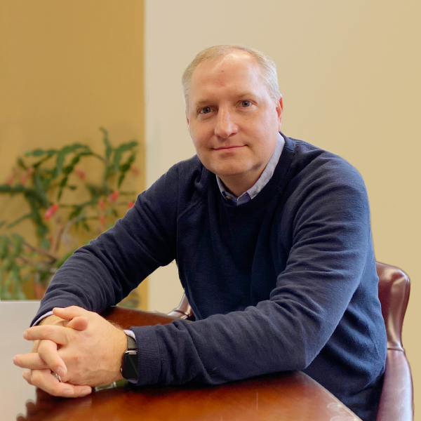 Mike Cahoon sitting at table, hands crossed, wearing dark blue sweater